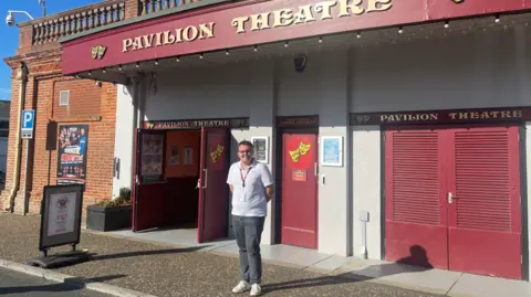 A man in a white polo shirt and grey jeans stands on a pavement outside the entrance to a theatre on a sunny day. Behind him are three sets of red doors and above his head the words Pavilion Theatre across the building in gold on red. The comedy/tragedy theatre masks are on the doors in yellow. 