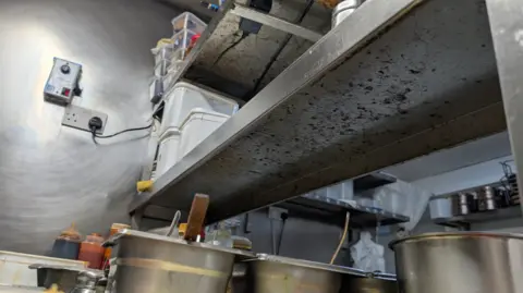 Oxford City Council A close up shot of the underside of a metallic shelf in a typical-looking small restaurant kitchen. Mould, appearing in a spread of dark blots, can be seen across the bottom of the shelf. Nearby there are bottled sauces and pans. 