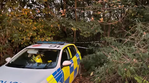 A police car parked in woodland. There is police tape across trees in the background.