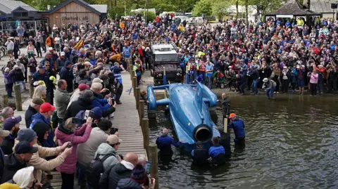 Getty Images Bluebird is gently lowered into the water backwards. Hundreds of people are gathered closely by and taking pictures on their mobile phones.
