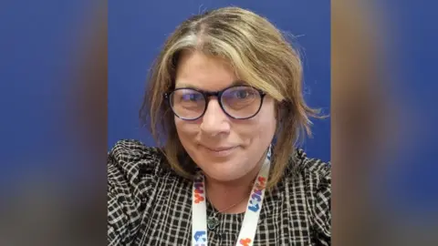 Nicky Twemlow A selfie of a woman with neck-length dark blonde hair and glasses. She is wearing a black and white check top and has a lanyard round her neck marked "YMCA".