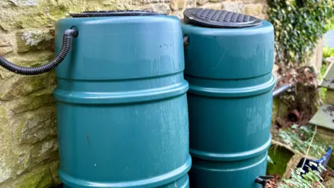 Getty Images Two plastic green water barrels against a stone wall linked by black hoses. 