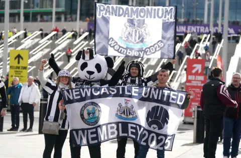 Owen Humphreys/PA Media A group of people, including someone dressed as a panda, hold a Newcastle United Wembley 2025 flag outside Wembley. Another flag says "pandamonium".