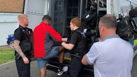 Police escorting a man into a police van. He is wearing trainers, shorts and a red jumper.