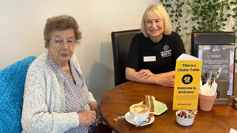 Two ladies sit at a table. An older lady who's wearing glasses is dressed in a blue shirt with white polkadots and a white and grey cardigan. In front of her there's a slide of cake and a coffee. Next to her there's a slightly younger lady wearing a volunteer shirt who smiles adoringly at her. On the table there is a triangular sign that says 'This is a Chatty Table".