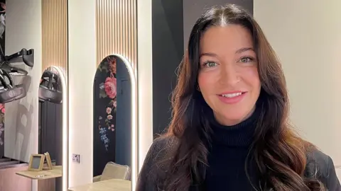 Salon owner Nat Cardno with long dark brown hair wearing a black polo neck jumper and blazer. She is standing in a hairdressers with mirrors and cream chairs in the background. She is smiling. 