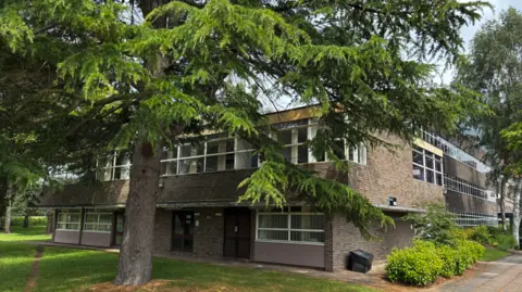 A brick building with large windows stands behind a big tree, with grass and a path in front.