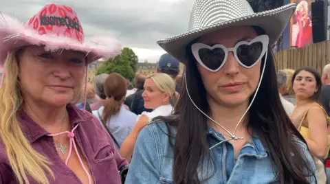 Rina standing on the right wearing a denim shirt, a disco ball cowboy hat, and white framed sunglasses shaped like a heart. Beside her is her aunt Stella, wearing a pink jumpsuit and pink cowboy hat decorated with lips and a fluffy rim. Behind them you can see the main stage and a large crowd of people.