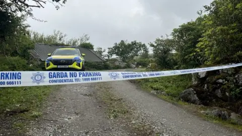 RTÉ The garda (Irish police) cordon at the scene of a fatal house fire at Gleann Mhic Mhuireann, County Galway. A yellow and blue garda car is parked on a rural country road, lined by trees and shrubs. A corrogated metal roof of a stone building is partially visible behind the car.