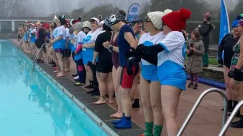 An image of a group of people dressed in swimming costumes with blue and white t-shirts on top and beanies and socks. They are preparing to jump into an outdoor pool. Some are wearing festive hats.