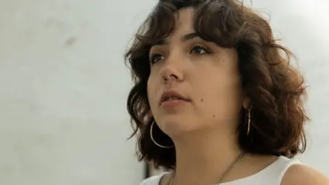 Riki Auton A woman with brown short curly hair wearing gold hoop earrings and a white vest top. 