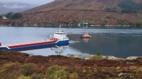 A red, blue and white coloured cargo ship close to shore with a lifeboat positioned behind it. There are white walled houses on the opposite shore.