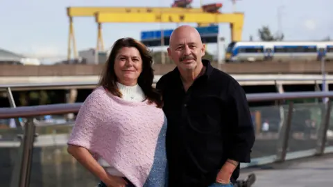 A woman in a pink top and a man in a black shirt. Yellow Harland & Wolff cranes are in the background.