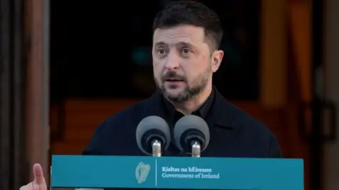 Zelensky - a man with short brown hair and band, stood in front of a microphone that reads Government of Ireland