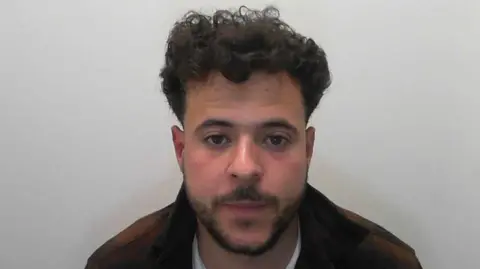 A police mugshot of Eid Anwar Fathi Najjar. A younger man with dark, curly hair, and a dark beard, wearing a brown jacket and white t-shirt.