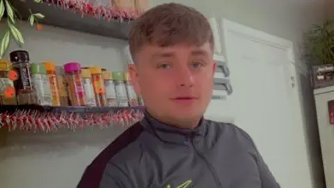 Family handout via West Yorkshire Police A man with brown hair and a brushed down fringe. He is wearing a grey zip-up tracksuit top and appears to be stood in a kitchen environment. 