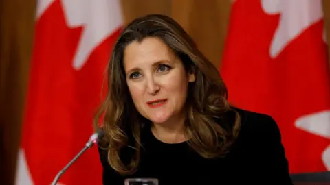 Reuters Chrystia Freeland, mengenakan atasan berwarna gelap, berbicara melalui mikrofon dengan latar belakang bendera Kanada. 