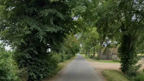 Google Gote Lane is a narrow, rural road surrounded by trees and shrubs. There is a small, old single-storey building to one side and a dusty track