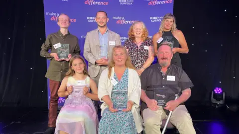 Seth Spain/BBC Left to right: James de Mierre, Toby Lindsay-White, Kathy Cummings, Tina Ashford, with Harli Dixon, Paula Baker and Paul Berryman in the front row. they sit on a stage in front of a banner which says 'make a difference' and each hold a glass trophy