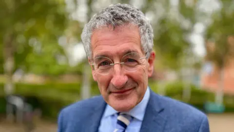 Martin Giles/BBC A man smiles at the camera. He has grey hair, glasses and is wearing a blue suit with a lighter blue shirt underneath as well as a cream and blue striped tie. 