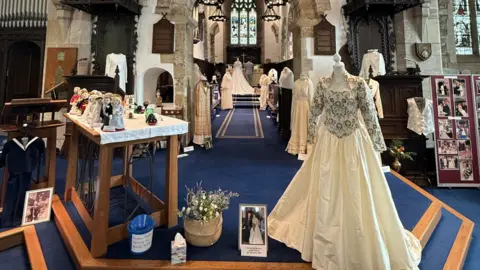An embroidered wedding dress stands on a mannequin with several others in the background. The dress is a centrepiece in the middle of St Andrew's Church with other wedding-themed items on display in an exhibition. 