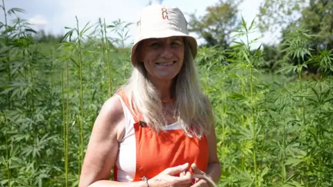Shaun Whitmore/BBC A woman smiles at the camera as she stands in a field where green hemp is growing. She wears a cream bucket hat with her long blonde hair flowing underneath it. She is also wearing a white vest top with an orange overall over the top.