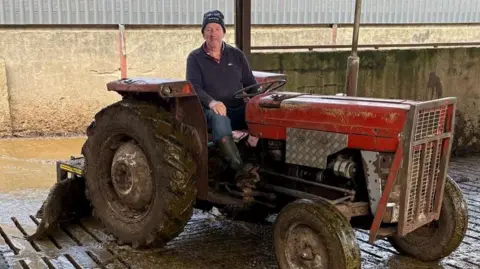 Brian Matthews A man is sitting on a red tractor inside a cattle shed. He is wearing a navy jumper, blue jeans, wellington boots and a navy beanie hat. 