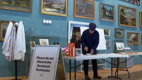 Cathy Minton/BBC A man in a blue flat cap makes crisp sandwiches at white table in an art gallery. In front of him is an A-board that reads 'Please do not disturb the sandwich technician'.