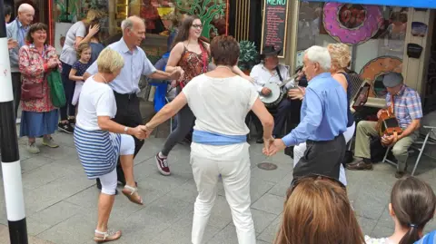 Getty Images Miembros del público tomados de la mano y bailando en un círculo con la música que una banda está tocando junto a ellos. Los rodean de ellos están mirando otras personas. Los miembros de la banda en Shot son un hombre mayor con un sombrero negro y una camisa blanca con pantalones negros tocando un banjo, y un hombre con una camisa a cuadros plana, zapatos marrones y pantalones verdes tocando un acordeón. Frente a ellos hay un anillo de seis personas, incluidas cuatro mujeres y dos hombres. Una mujer tiene cabello rubio y lleva una tapa blanca, pantalones y sandalias con una chaqueta azul y blanca alrededor de su cintura, la siguiente mujer tiene cabello corto y castaño y también lleva ropa blanca con una prenda azul envuelta alrededor de su cintura. El hombre que está a su lado tiene una camisa azul y pantalones grises con el pelo corto gris, detrás de él, se puede ver la parte posterior de una mujer con cabello rubio corto, pantalones blancos y una parte superior azul marino. Luego hay una chica más joven con cabello largo y castaño, gafas, jeans negros y jeans grises. Un hombre calvo con pantalones negros está bailando a su lado.