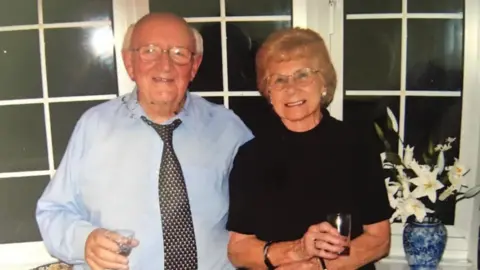 Chelsea Boyce A man is wearing a blue shirt and a black and white polka dot, stood next to a woman wearing a black top. Both of them are smiling at the camera.