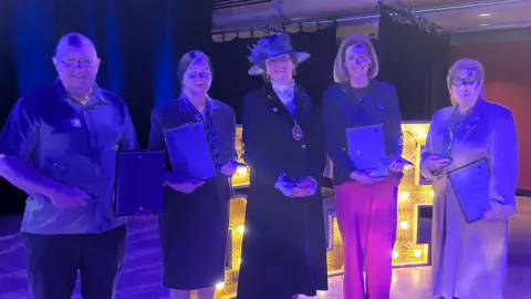 Jon Wright/BBC Ian Walters, Helen Fairweather, Yvonne Gilchrist-Mason, Hannah Tuckwell and Katherine Ahluwalia  - stand holding glass trophies and certificates. The blue stage lighting has washed over their faces.