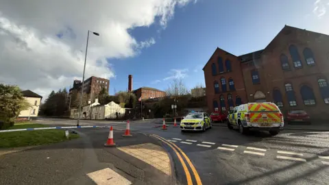 Police vehicles guard a cordon at the bottom of a hill. There is a large mill structure in the background which appears to be derelict. Traffic cones and police tape also mark the perimeter of the cordon.