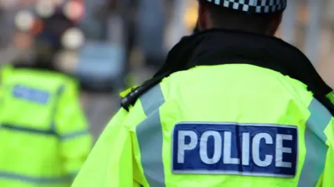 A stock shot of two police officers, one close to the camera, both with their back to the lens, wearing high-vis jackets. The luminous yellow jackets have the word "Police" written on them.