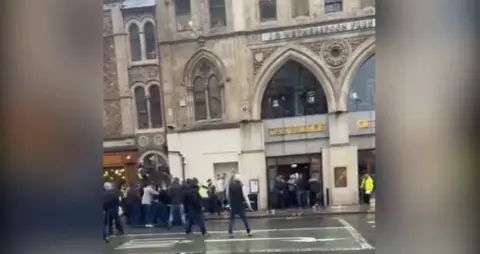 A group of people standing on the road outside the Prince of Wales pub in Cardiff. A police officer on a horse can be seen in the left hand side of the screengrab while around a dozen people can be seen in the middle of the shot, facing away from the camera. At least two police officers can be seen in the group.