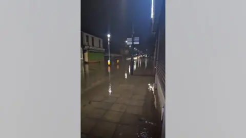 I Am Bored North East Chester Road following the burst water main. Water has flooded the road and pavement. Light from traffic lights is reflected in the water.