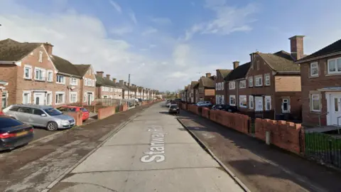 Google Maps street view of Stanway Road, Gloucester. Red brick houses line both sides of the street and cars can be seen parked on driveways. 