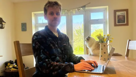 A young man sitting down with his hands on a laptop's keyboard. He is wearing a blue fleece and in the background there is a sliding door and a cat. 