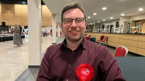 Amy Holmes/BBC A man with glasses wearing a red rosette that reads “I’m Voting Labour” is seated at a table inside a large indoor hall. The space has a row of bar taps along a counter to the right, with red chairs and tables arranged around the room.