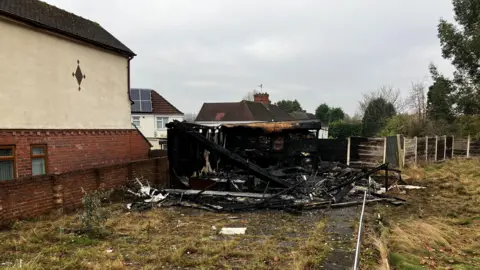 The charred remains of a building, next to a wall. There are houses in the background and a house on the other side of the wall. There are fence panels - some of which are also burned - and in the foreground are some paving slabs which are overgrown with vegetation.