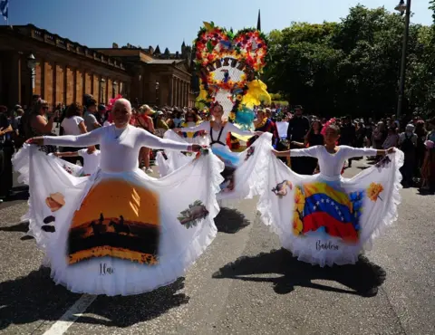 Parade performers in white dresses display wide skirts printed with colourful images and a Venezuelan flag, while processing along a city street. A large floral display and crowds of spectators follow behind on a sunny day.