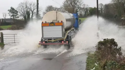 A milk tanker splashing through a flooded road. There are trees in the background. 
