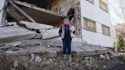 Angelina stands in front of the ruins of the school clutching a teddy