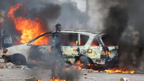 Reuters A protester stands in front of the shell of a car that is on fire. Flames and smoke billow out of the vehicle. 