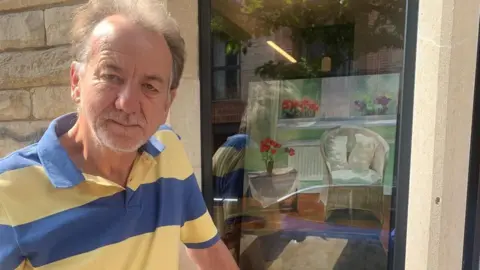 David Finch standing in front of a window where his painting is displayed. He has short fair hair and facial hair. The painting depicts a chair and a table with a bunch of flowers in a vase. David is looking at the camera and smiling