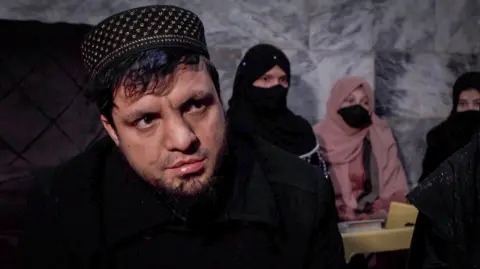 Hamid sits in front of some girls in a classroom. 