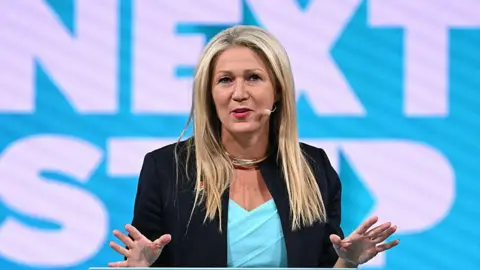 Getty Images Laura Anne Jones wearing a black jacket and blue top, standing at a podium with her hands raised. She has a white and blue backdrop behind her and is wearing a microphone.