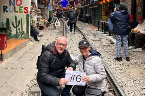 Tony and Tracy Raine Hanoi's famous railway street is a narrow 400 metre residential stretch where trains pass inches from cafes. In the foreground Tony has a black puffer coat on, Tracy has a light silver puffer jacket with a black baseball cap. The couple are holding a number 6 on an A4 sheet of paper.