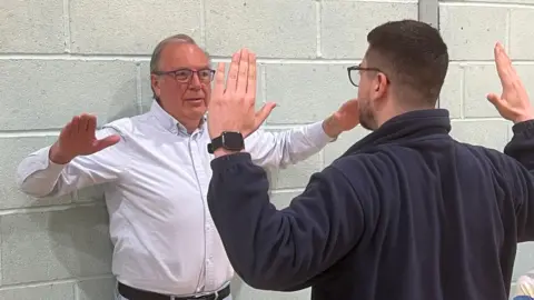Martin, wearing a shirt and jeans, is stood with his back against a wall. His arms are raised to be almost level with his shoulders with his elbows bent at 90 degrees and his palms in front of him. He is mirroring the pose of a physiotherapist, who is stood in the same position with his back to the camera.