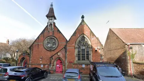 Google Street View Beverley Minster Parish Hall before the bell tower was removed. There are cars parked in front of the red brick building.