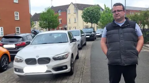 Daniel Adams Councillor Daniel Adams standing on the pavement in Redhill Way, North Swindon next to a line of cars parked on the pavement with trees and houses in the background.